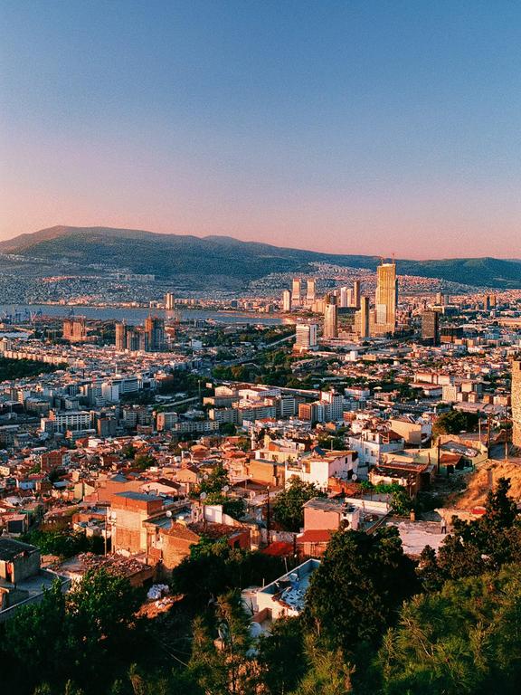 A city with a mix of old and new buildings stretches towards a body of water and mountains under a clear sky.