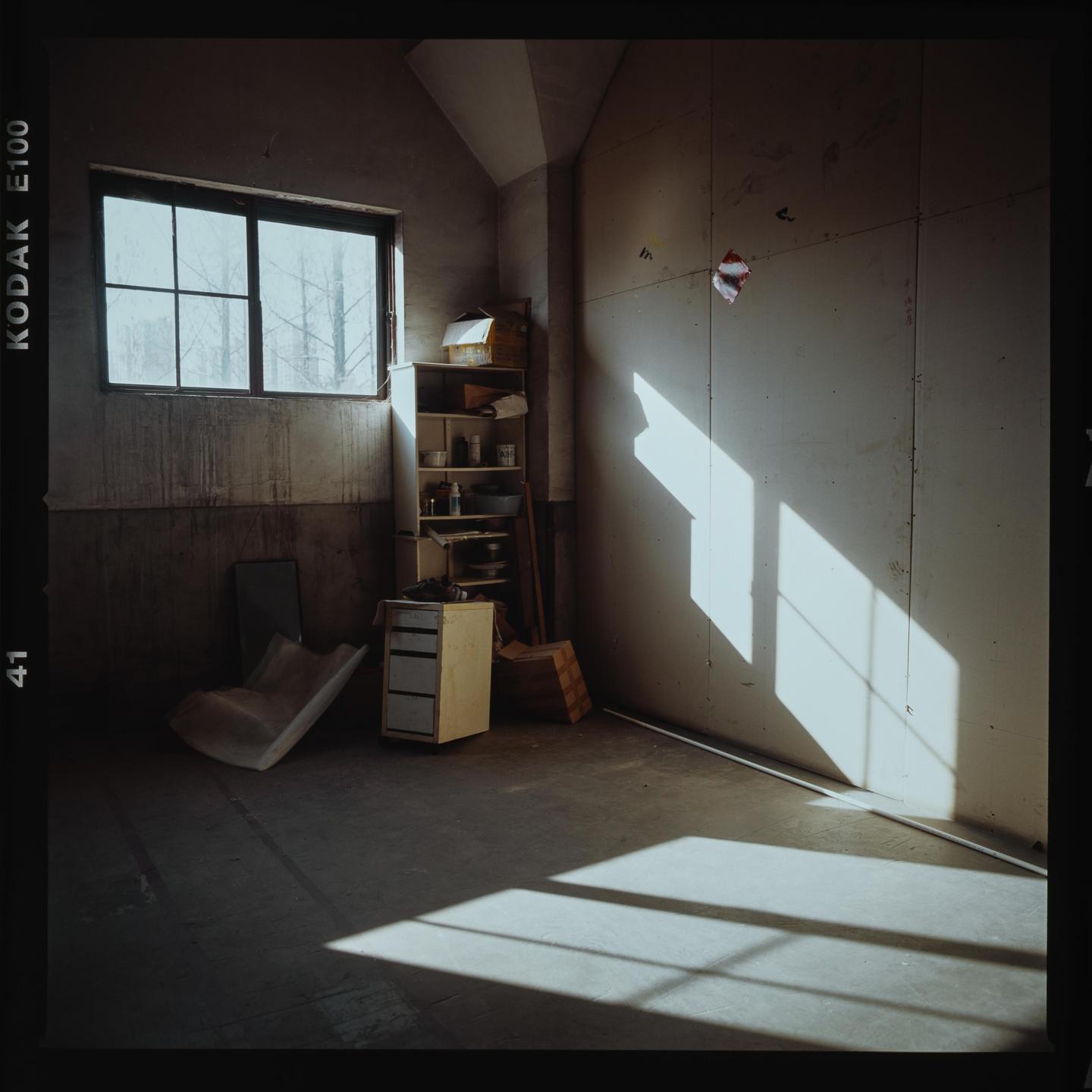 Sunlight streams through a window into a sparsely furnished room with concrete floors and a wooden shelf.