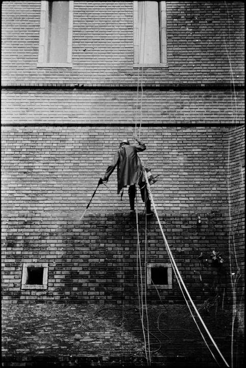 A man in a protective suit is suspended by ropes, power washing a brick building.