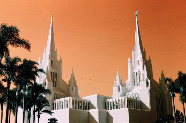 A white temple with multiple spires stands against an orange sky, framed by palm trees.