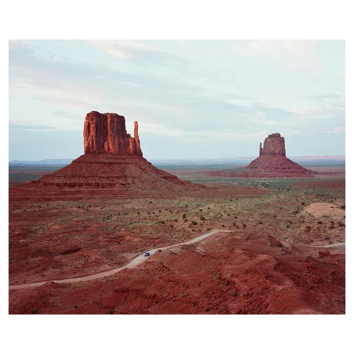 A small car drives on a dirt road through a desert landscape with two large rock formations under a pale sky.