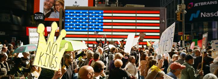 A crowd of people are protesting in Times Square at night, holding signs and umbrellas, with a large American flag display in the background.