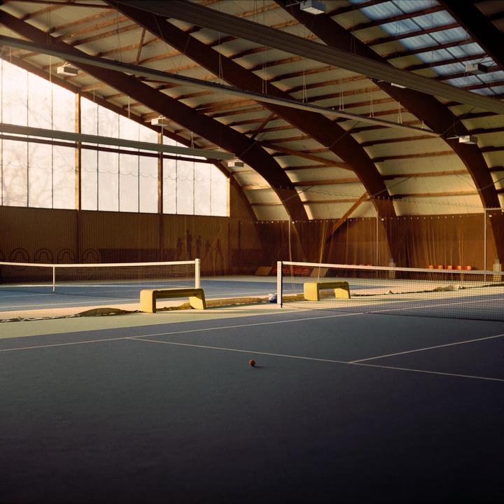 An indoor tennis court with a single tennis ball on the ground is illuminated by sunlight streaming through large windows.