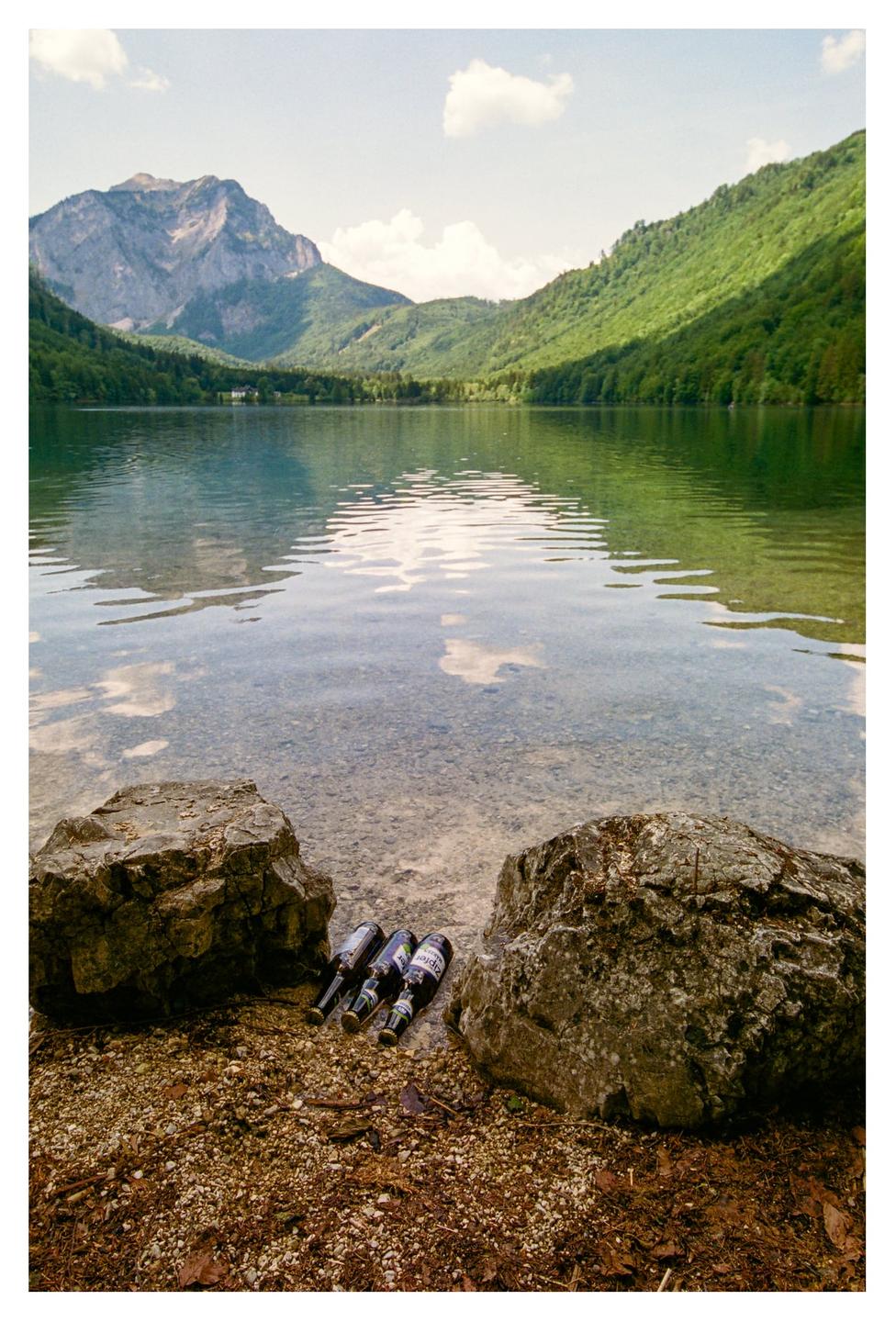 Three bottles rest on the rocky shore of a lake surrounded by mountains and forest.