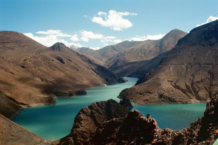 A turquoise river winds through a valley surrounded by arid mountains under a blue sky with scattered clouds.
