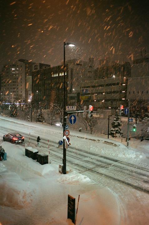 A snow-covered street at night is illuminated by streetlights and traffic lights, with buildings and trees in the background.