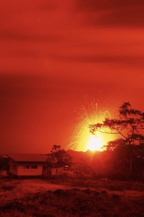 A house and trees are silhouetted against a bright orange sky with a volcanic eruption in the distance.