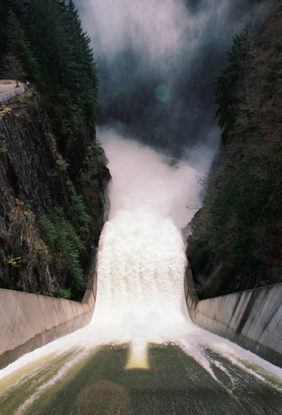 A large volume of water flows over a dam spillway, surrounded by steep, forested cliffs.