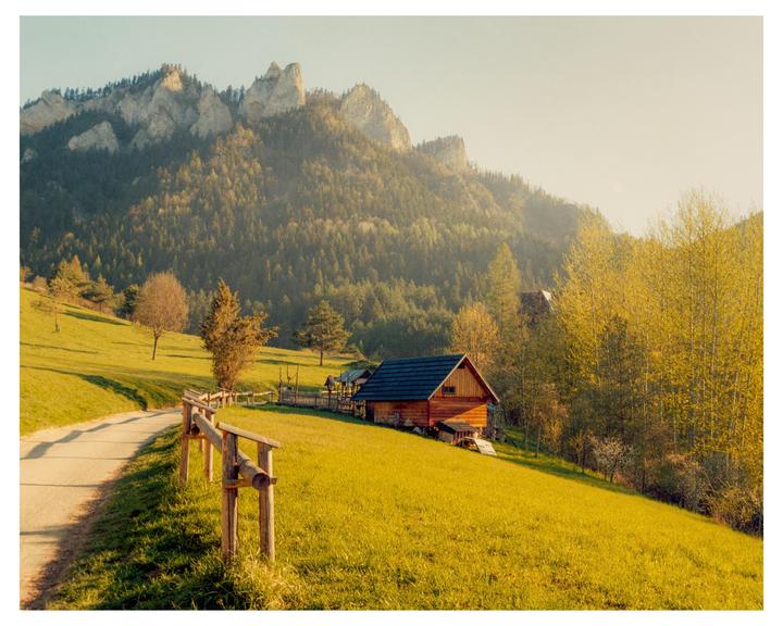 A wooden cabin sits on a grassy hillside with a mountain range in the background.