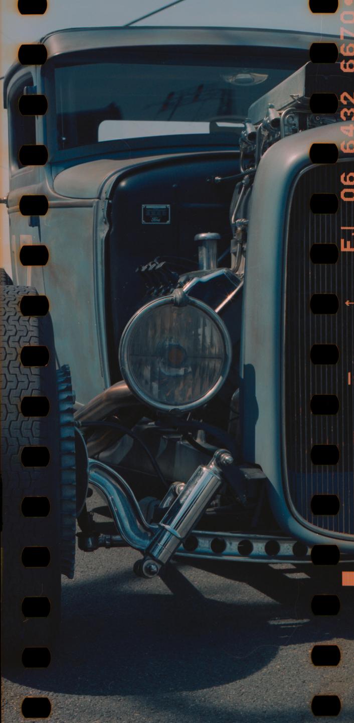 A close-up view of the front of a hot rod shows its headlight, grille, and exposed engine components.