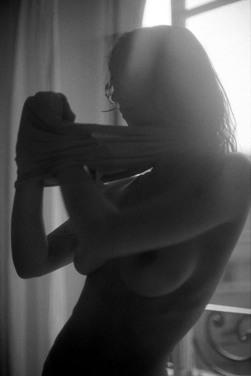 A woman is seen from the side, pulling a shirt over her head in a backlit room.