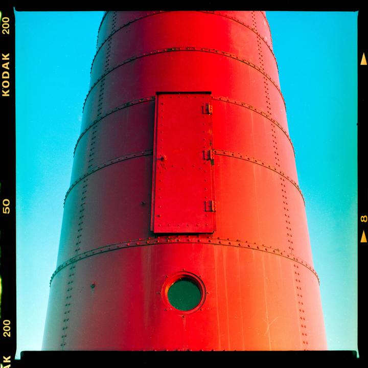 A red lighthouse with a door and a round window is set against a clear blue sky.