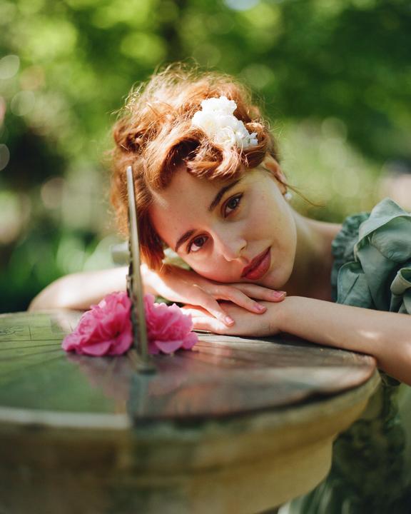 A woman with red hair and a white flower in her hair rests her head on her hands on a sundial with a pink flower.