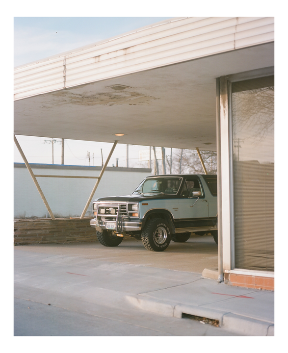 A light blue and black Ford Bronco is parked under a mid-century modern carport with a man in the driver's seat.