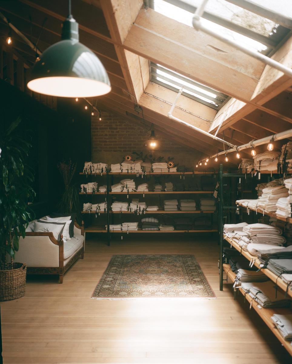 A large room with a high ceiling and skylights contains shelves of folded textiles, a sofa, and a patterned rug on a wooden floor.