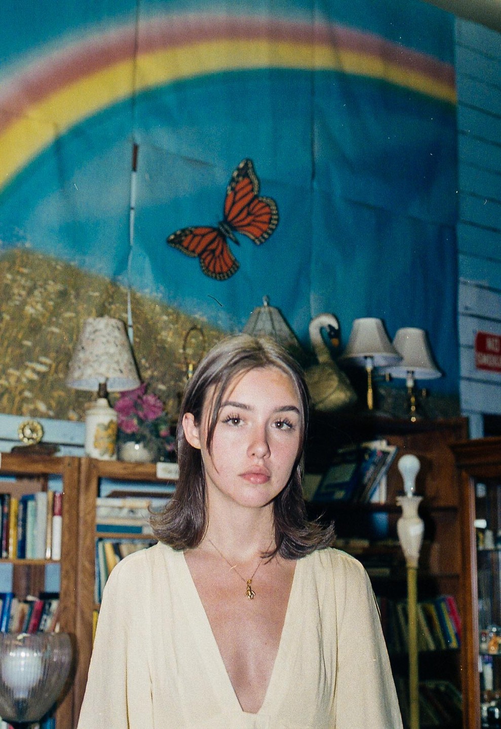A woman with a gold necklace stands in front of a blue wall with a rainbow and butterfly decoration, with bookshelves and lamps visible in the background.