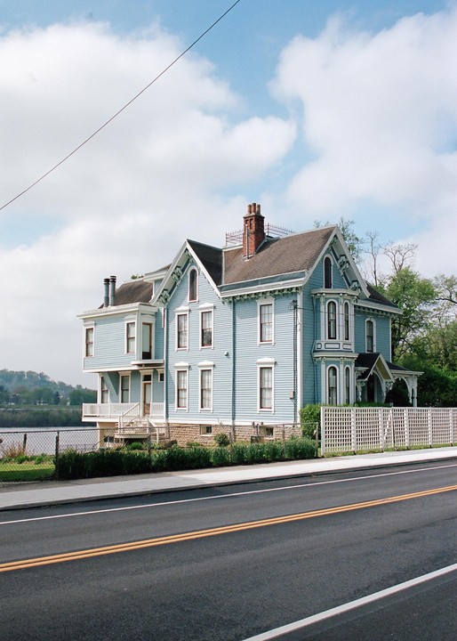 A stately blue house with a white fence stands beside a road, with a river and hills in the background under a cloudy sky.