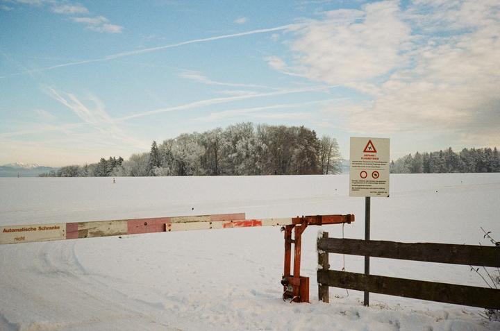 A barrier and a warning sign stand in a snow-covered field with a forest in the background.