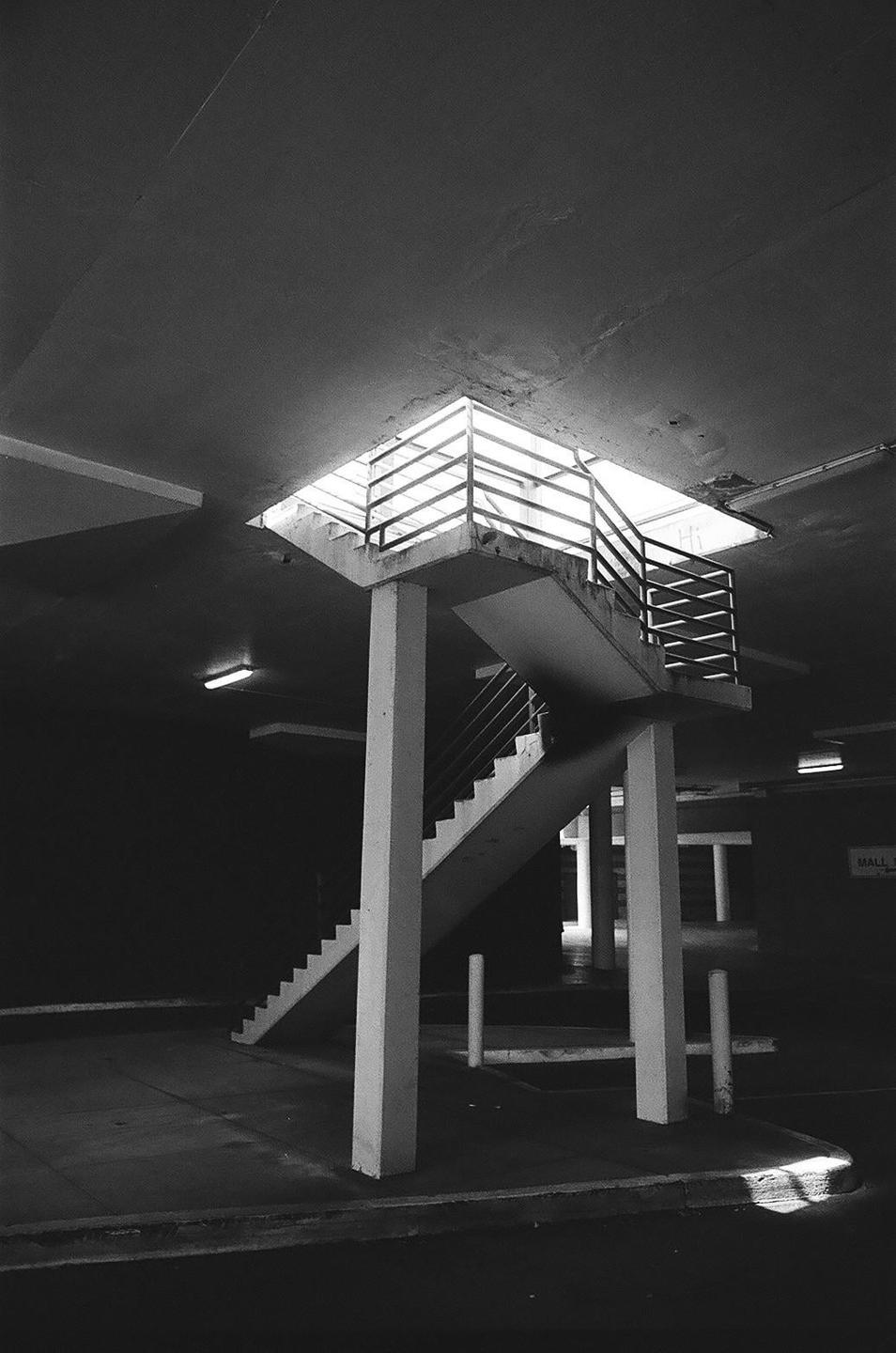 A concrete staircase with metal railings ascends towards a bright opening in a dark, enclosed space.