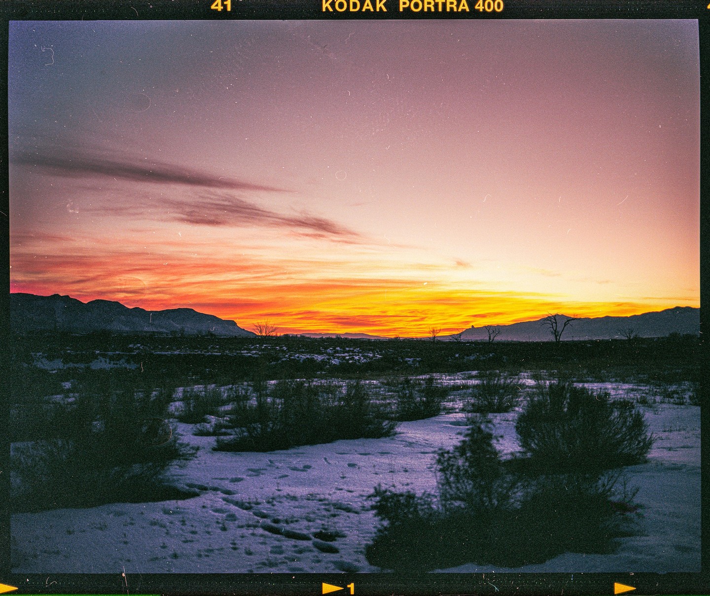 A vibrant sunset illuminates a snowy landscape with mountains and sparse vegetation under a colorful sky.