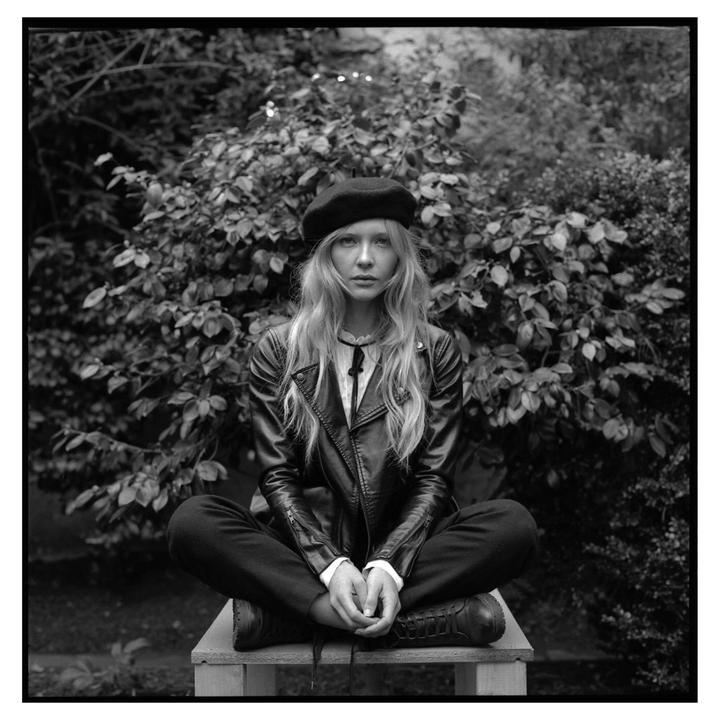 A woman wearing a beret and leather jacket sits cross-legged on a wooden bench in front of a leafy bush.