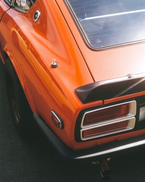 The rear of an orange Datsun car is visible, showing its taillights, spoiler, and part of the back window.