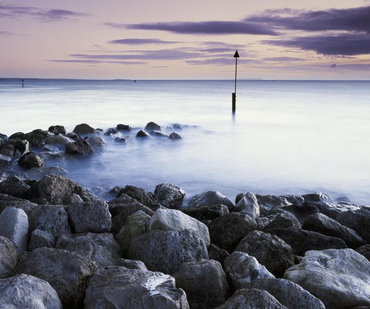 A rocky shore meets a calm sea under a sky with purple clouds.
