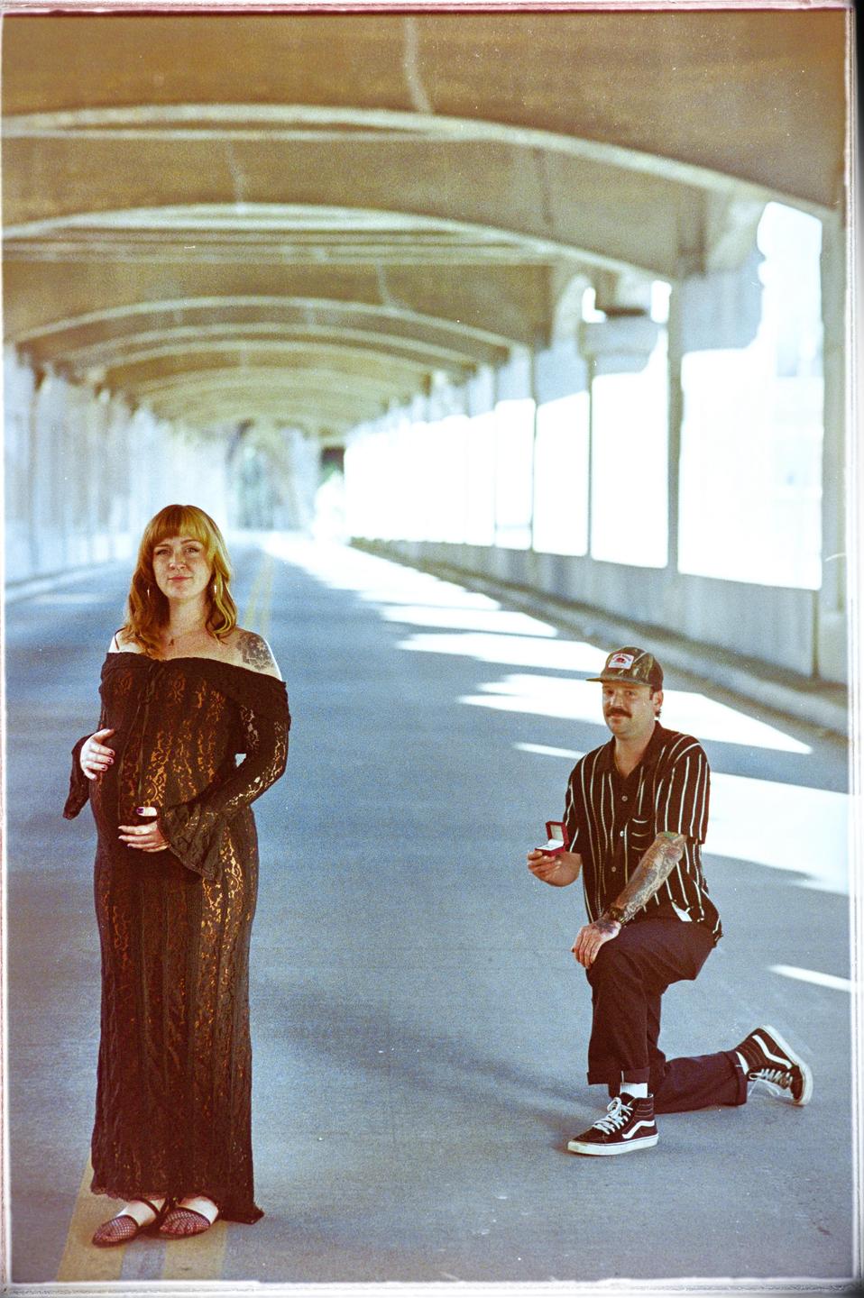 A pregnant woman stands next to a man kneeling with an engagement ring in a tunnel.