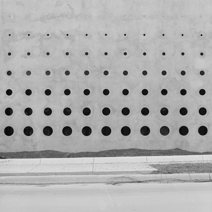 A concrete wall with rows of circular openings of varying sizes stands above a sidewalk and street.