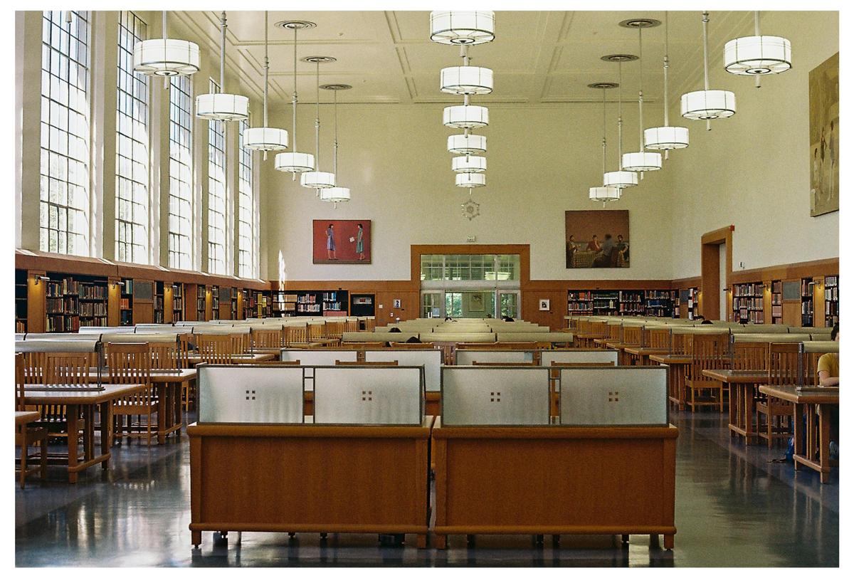 A large library reading room with tall windows, long wooden tables, and hanging circular lights.