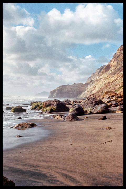 Waves crash on a rocky beach with cliffs in the background under a cloudy sky.