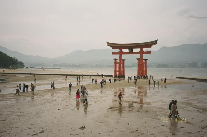 A large red torii gate stands in shallow water with many people walking on the wet sand around it, and mountains are visible in the hazy background.