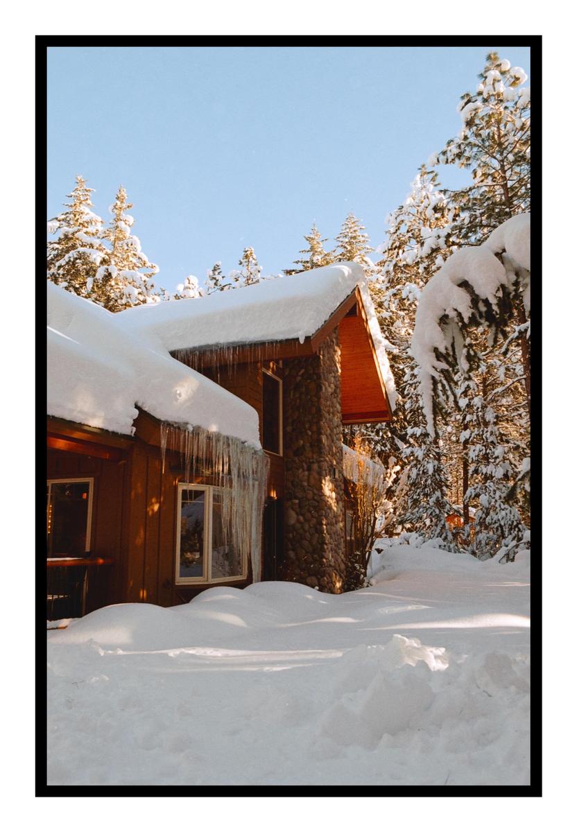 A snow-covered cabin with icicles hanging from the roof is surrounded by snow-laden pine trees under a clear sky.