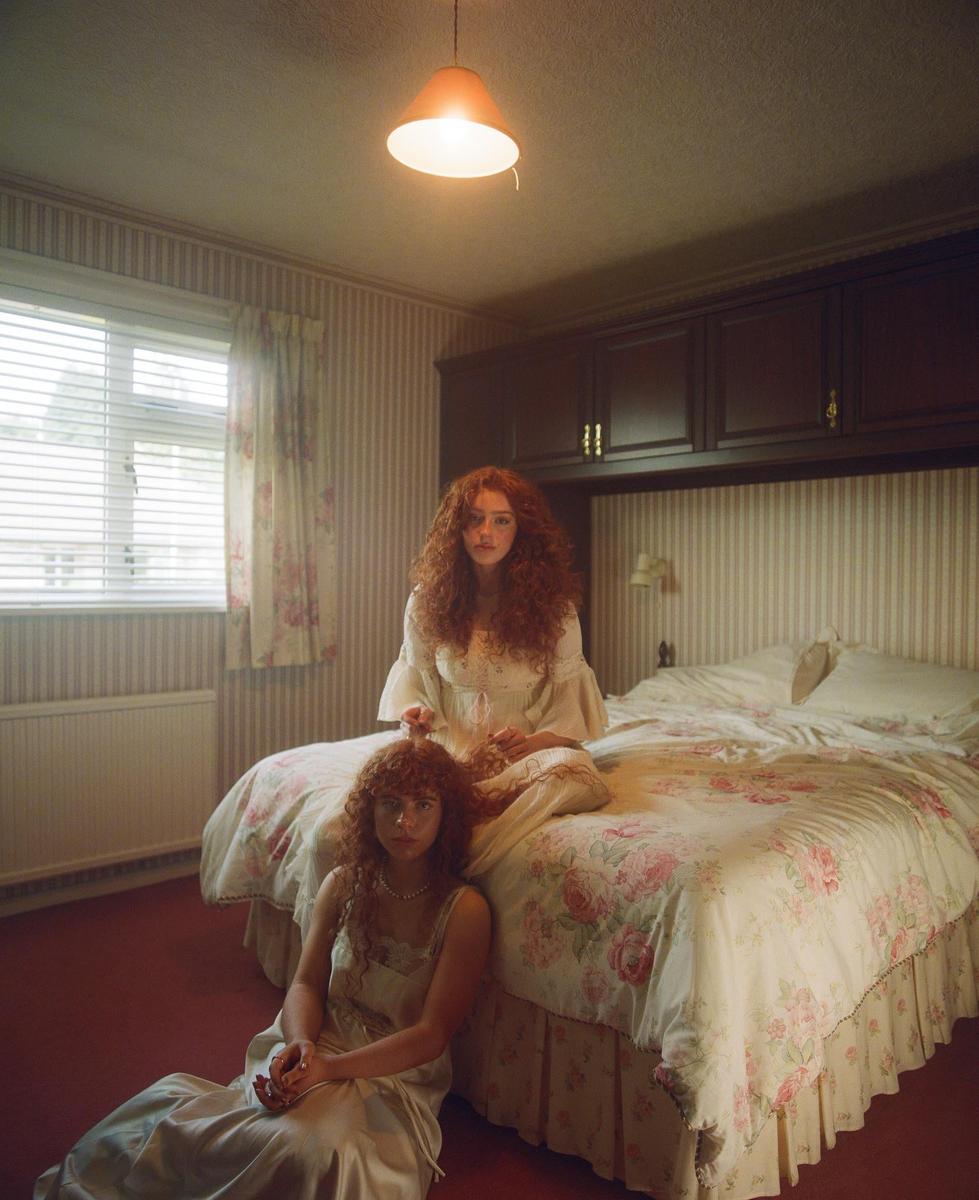 Two women with curly red hair are in a bedroom, one sitting on the bed and the other on the floor.