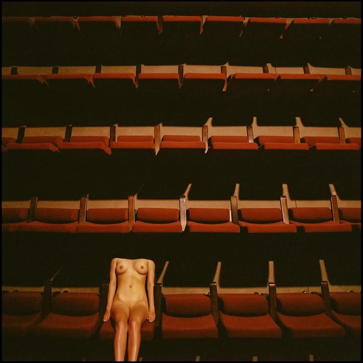 A nude woman sits in a theater with many rows of empty seats.