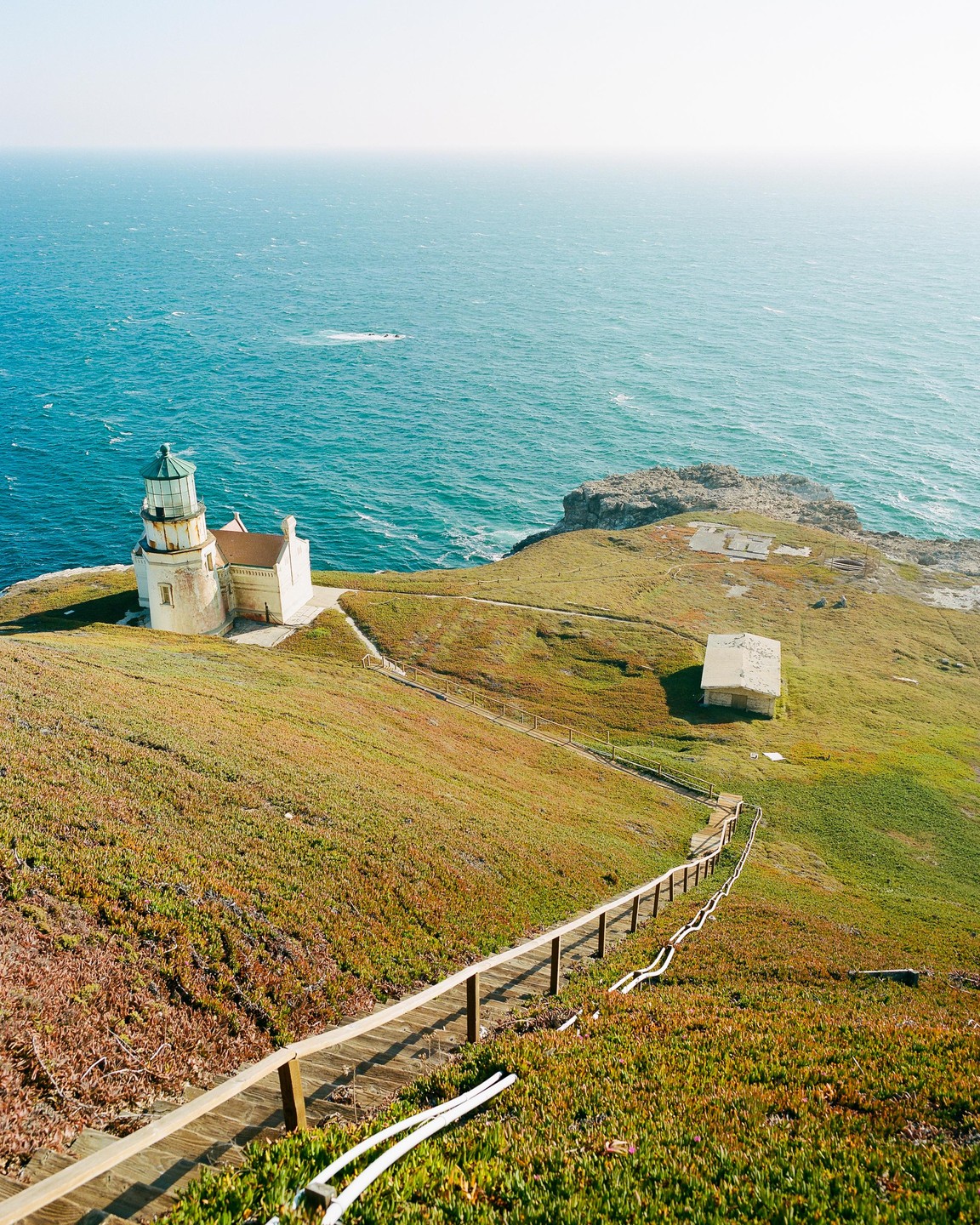 A lighthouse stands on a grassy cliff overlooking the ocean, with a long wooden staircase leading down the hillside.
