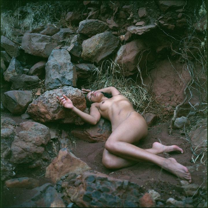 A nude woman lies on the ground among rocks and dry grass.