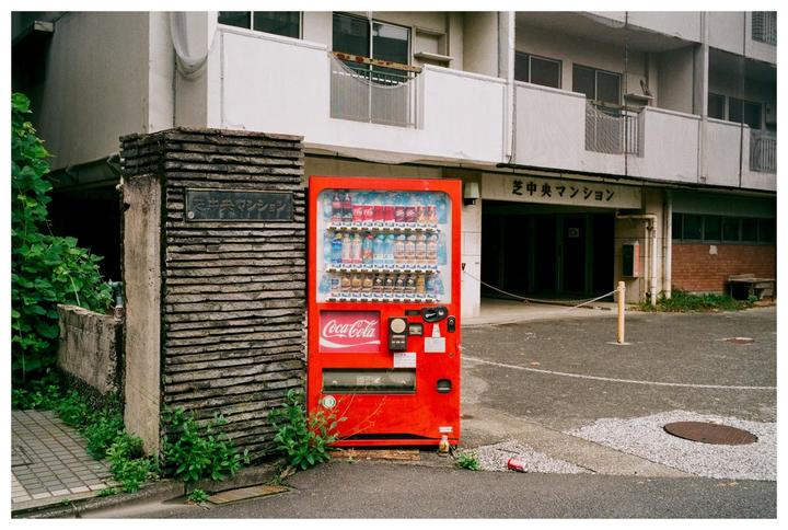 A red Coca-Cola vending machine stands next to a stone pillar with Japanese writing on it, in front of a building.