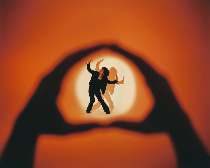 A woman with curly hair stands in a spotlight, framed by a large shadow of hands forming a circle.