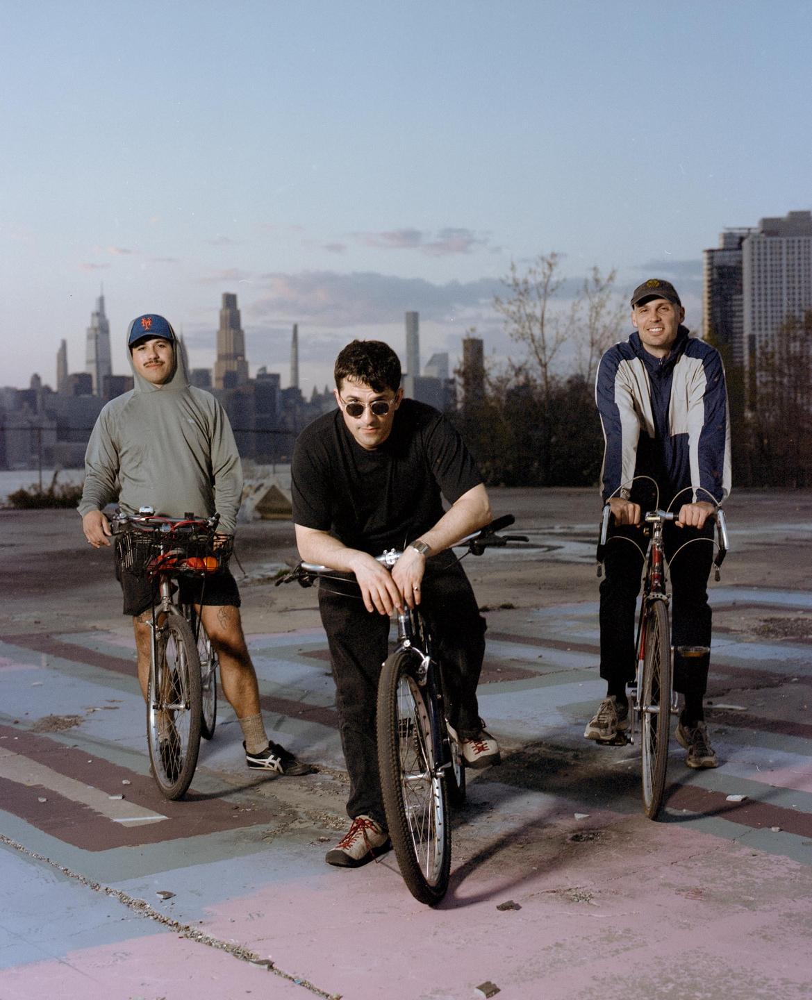 Three men pose with their bicycles in an empty lot with a city skyline in the background.