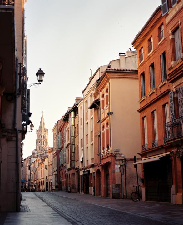 A narrow cobblestone street is lined with buildings and a church tower is visible in the distance.