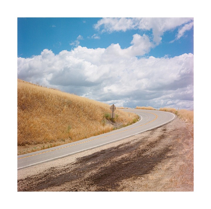 A winding road curves through a dry, grassy hill under a cloudy sky.