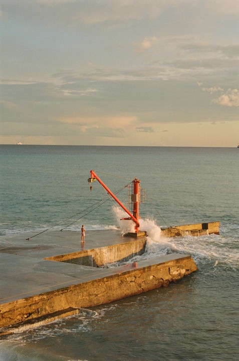 A man stands on a concrete pier next to a red crane as waves crash against the structure.