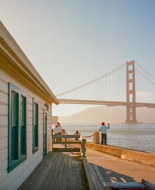 A person in a red hat takes a photo of the Golden Gate Bridge from a pier next to a white building with green trim.