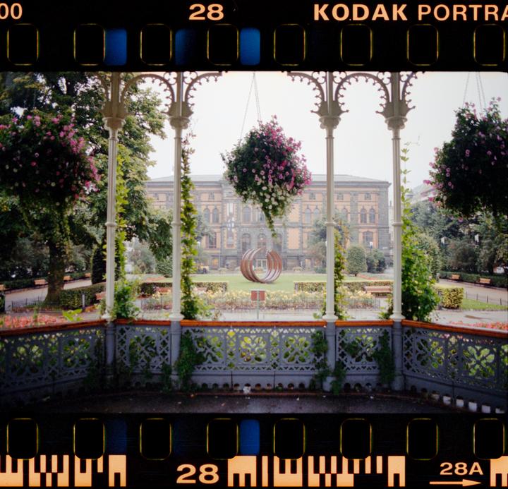 A park with hanging flower baskets and a building in the background is viewed from inside a gazebo.