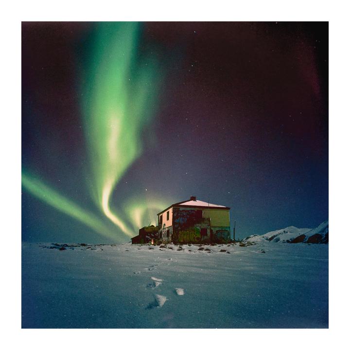 The northern lights glow above a house in a snowy landscape with mountains in the distance.