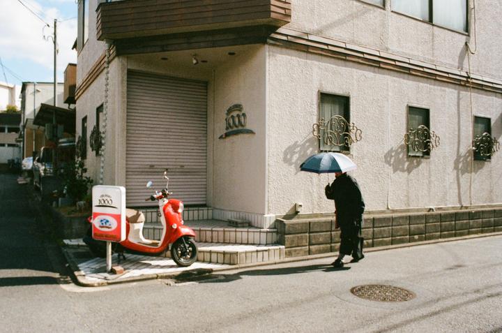A person holding an umbrella walks past a red scooter parked on a street next to a building.