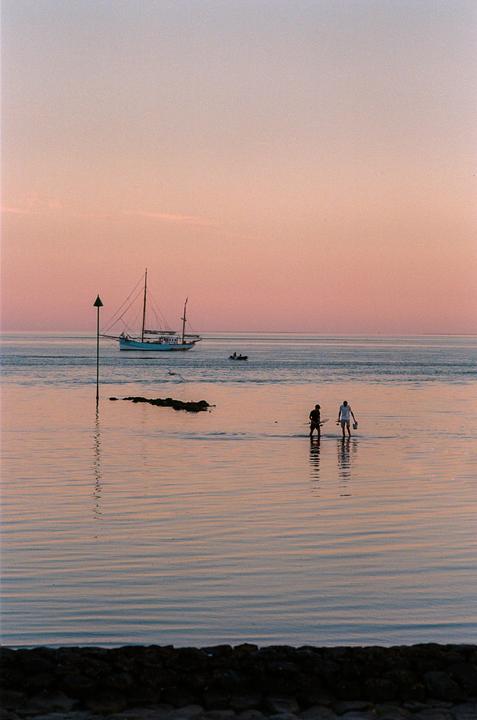 Two men wade in shallow water with a sailboat and a small motorboat in the background under a pink sky.