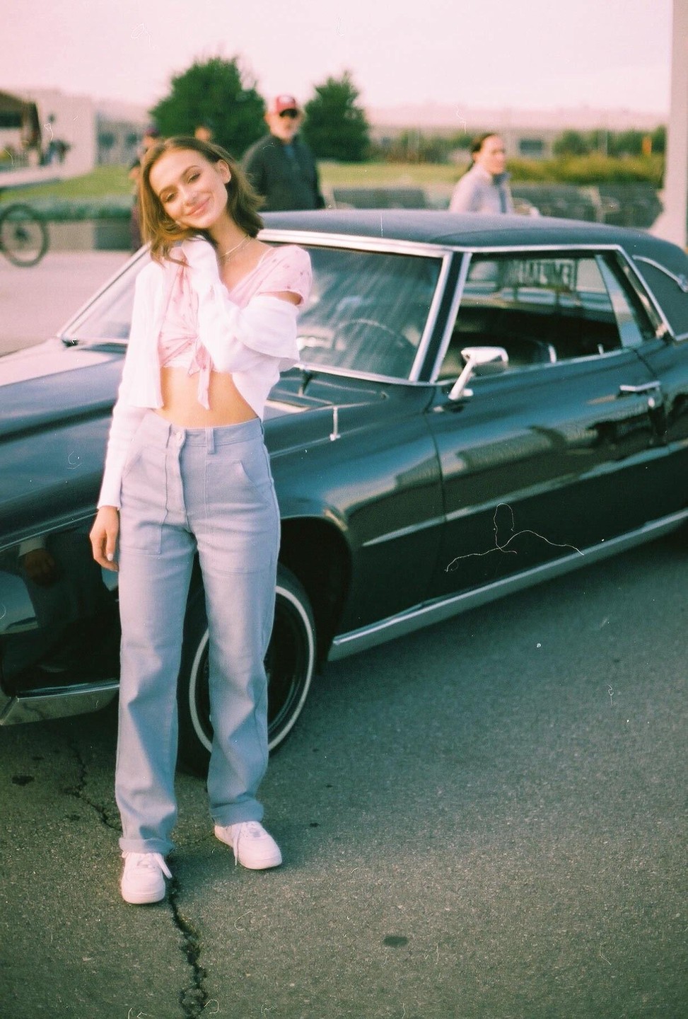 A young woman in a pink top and light blue pants smiles while posing next to a dark vintage car.