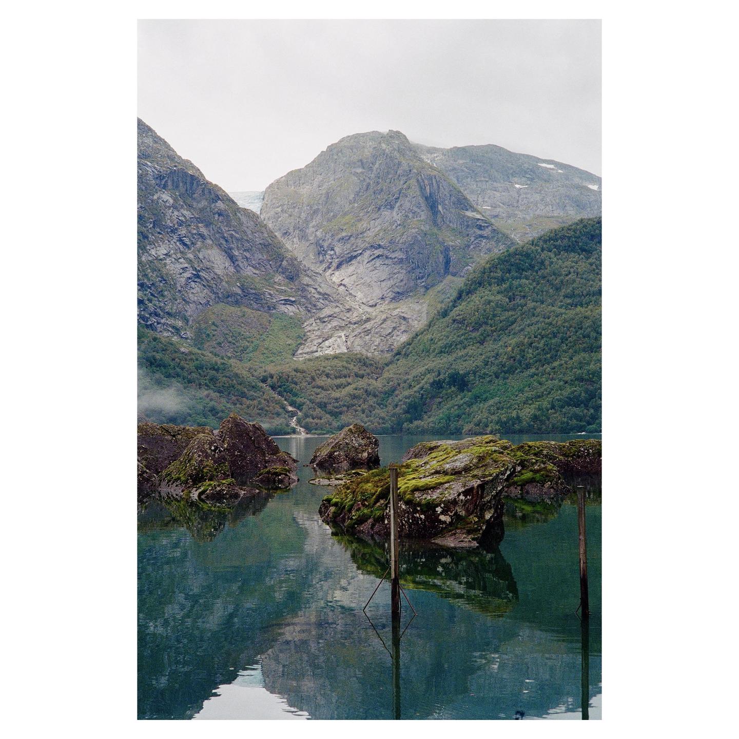 A body of water with mossy rocks and wooden posts reflects mountains and a cloudy sky.